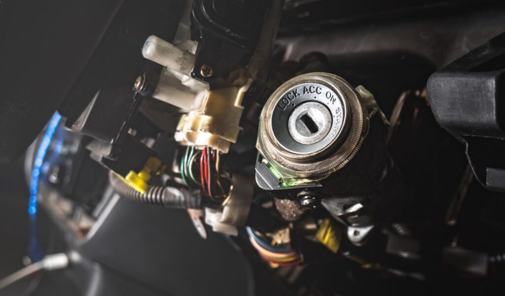 Exposed car ignition lock cylinder marked LOCK, ACC, ON and START with steering column wiring behind it