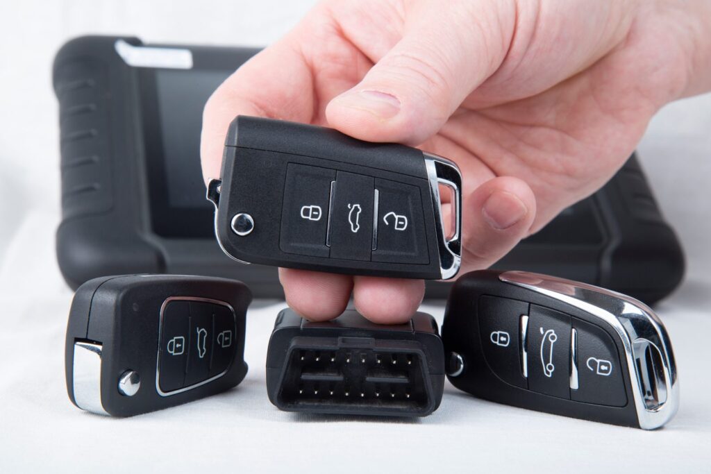 Hand holding a flip key fob above two other car remotes, an OBD connector and a key programming tablet